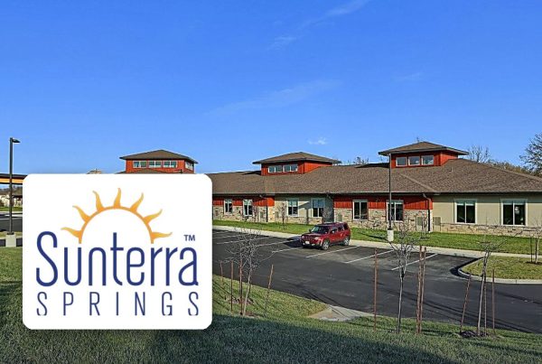 Exterior view of a single-story Sunterra Springs facility with a parking lot and blue sky; Sunterra Springs logo displayed in the foreground of the image.